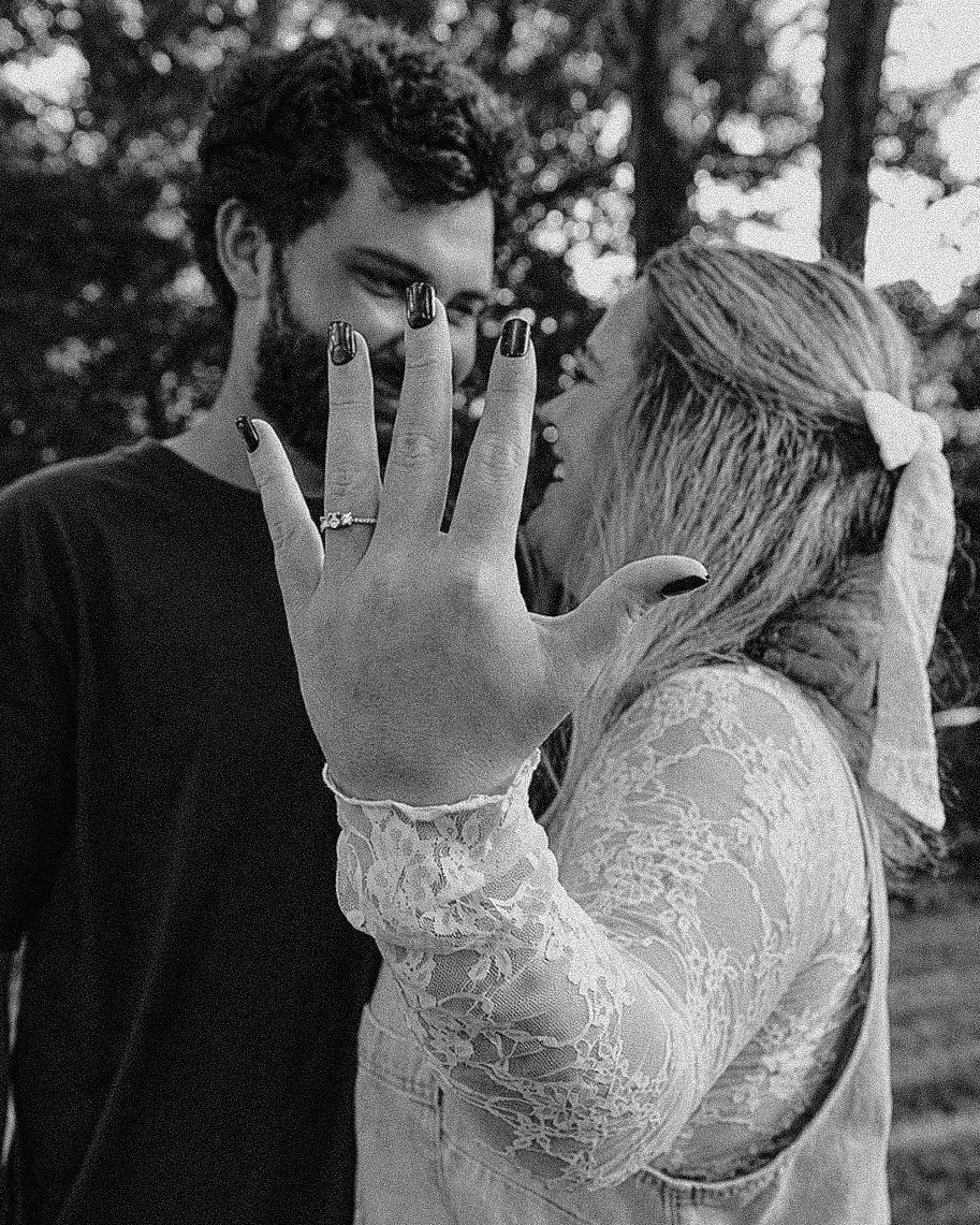 Black and white photo of a couple almost kissing, woman showing off her engagement ring to the camera.