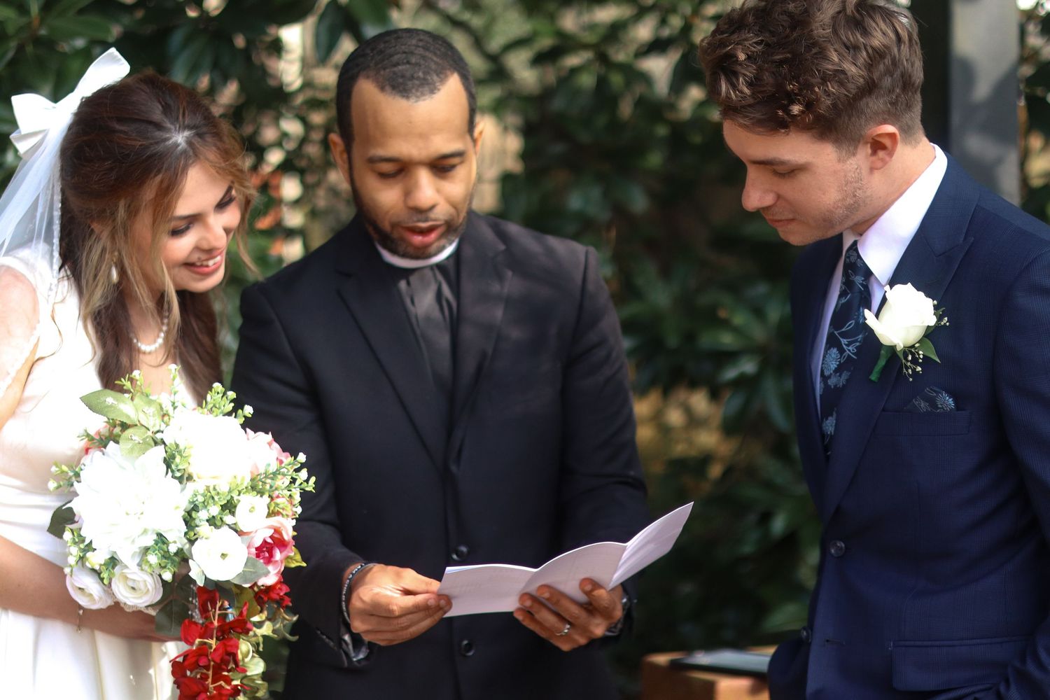 An outdoor wedding ceremony with a couple and officiant surrounded by greenery while reading from papers.