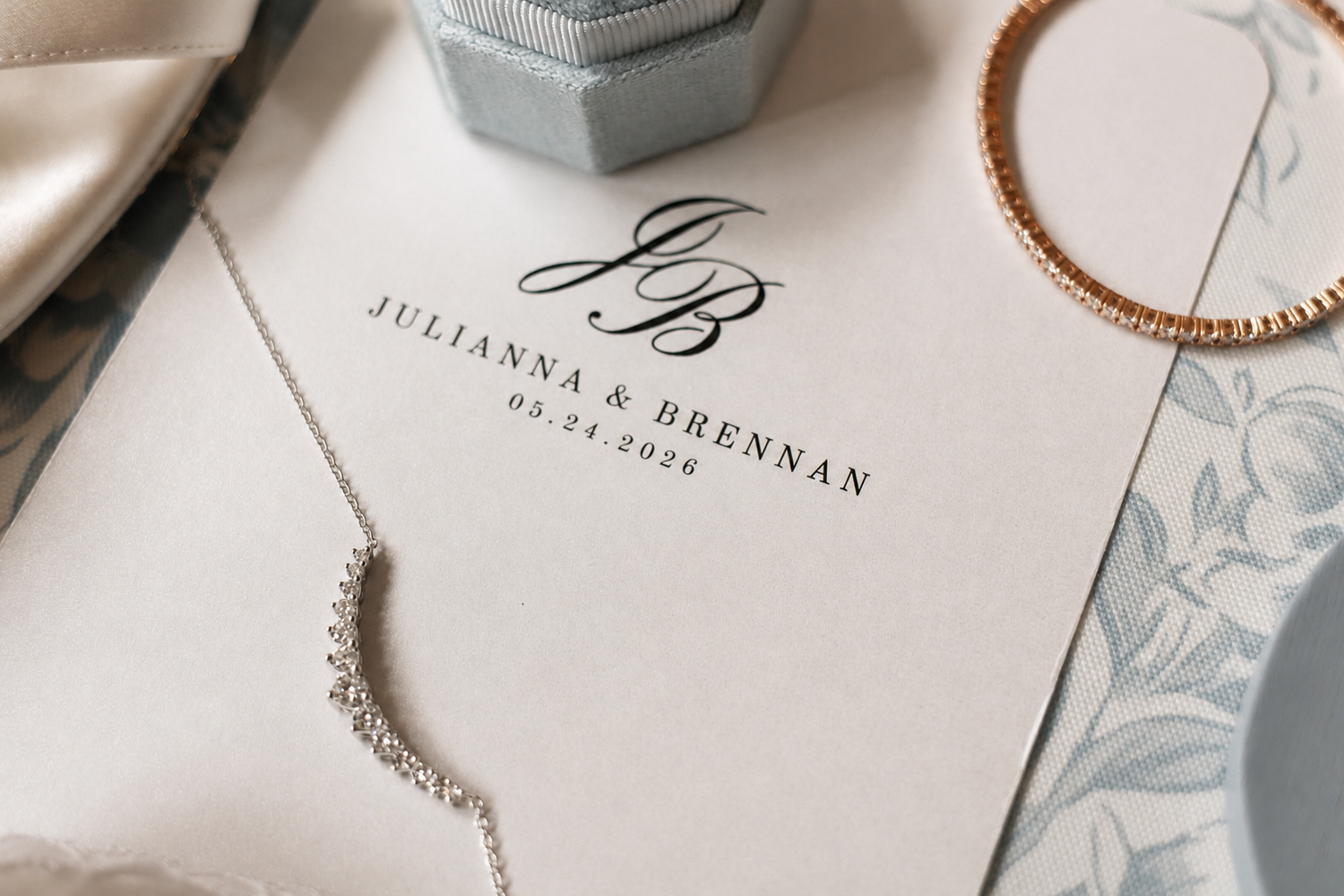 Elegant wedding stationery for Julianna and Brennan with monogram, beside a diamond necklace, gold bracelet, and jewelry box.