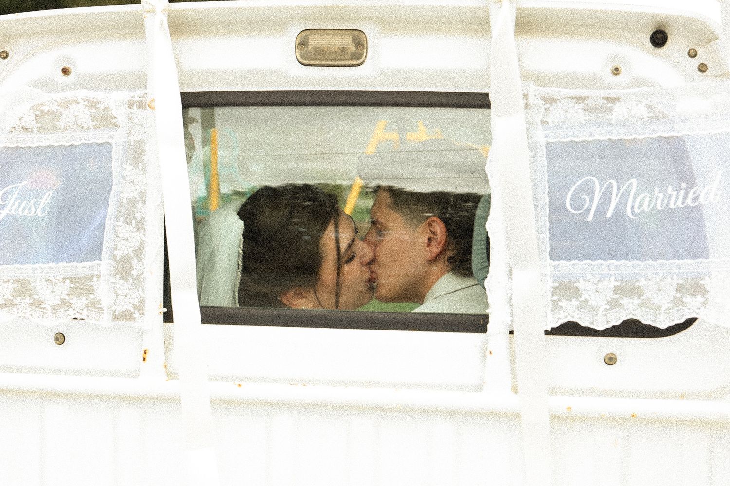 Newlywed couple kissing inside a white vehicle, seen through the rear window decorated with lace and a Just Married sign.