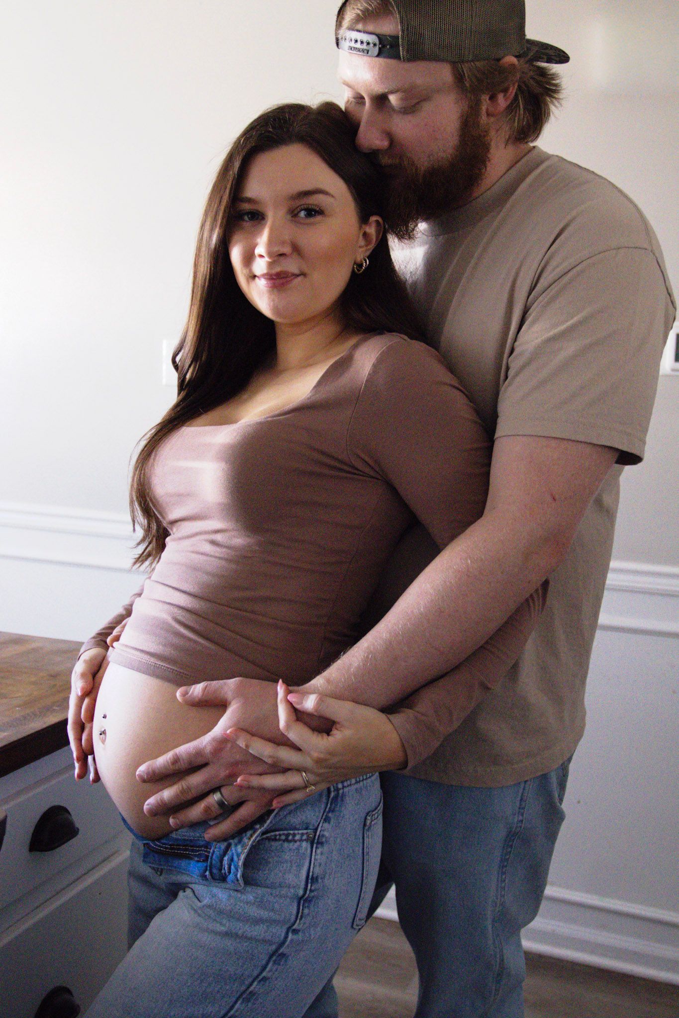 Pregnant woman in brown top and jeans poses with bearded man embracing her baby bump indoors.