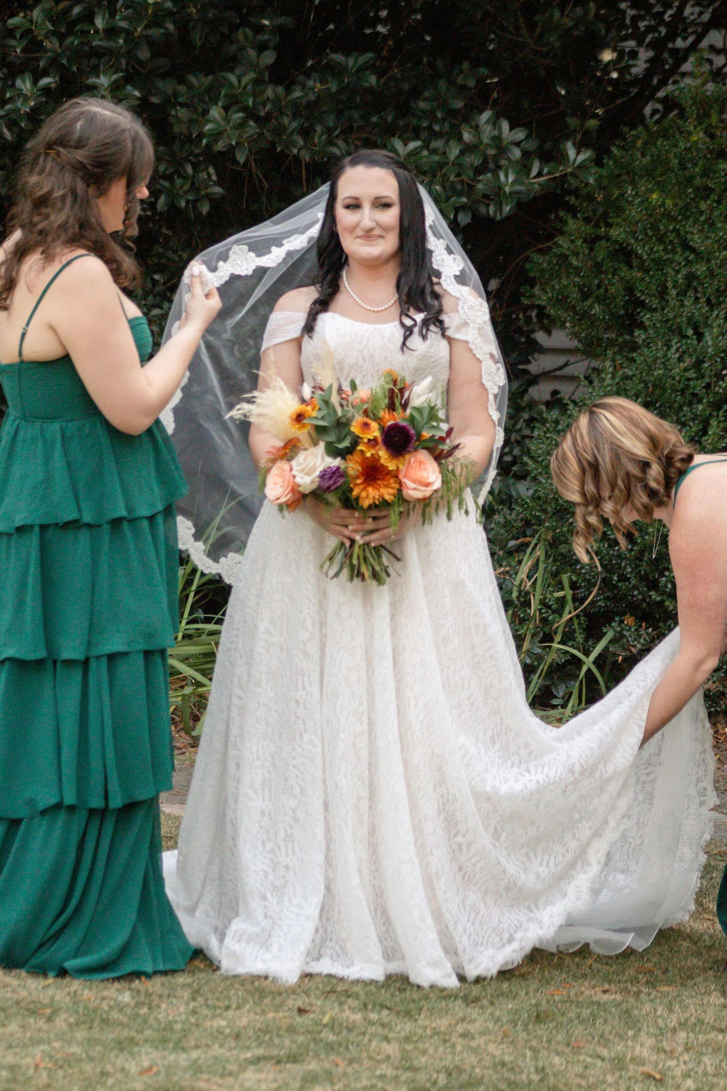 A wedding scene outdoors with attendants in green and white dresses helping with a lace gown and sunflower bouquet.