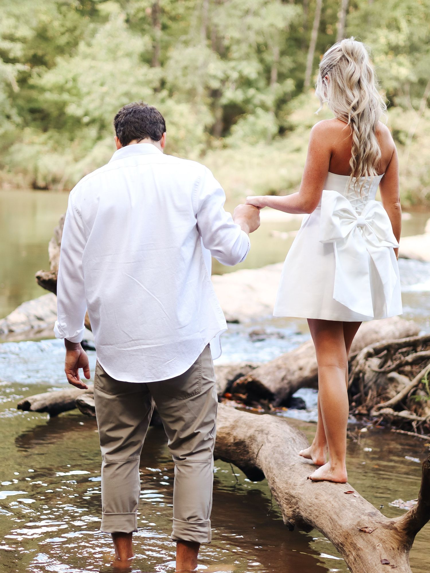A couple stands on a fallen log over a stream, with the woman wearing a short white dress with a bow.
