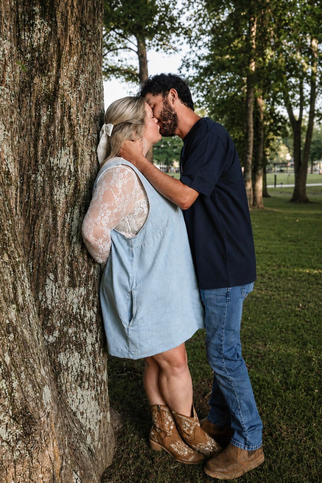 Couple sharing a romantic moment leaning against a tree in a park, woman in blue dress and boots.
