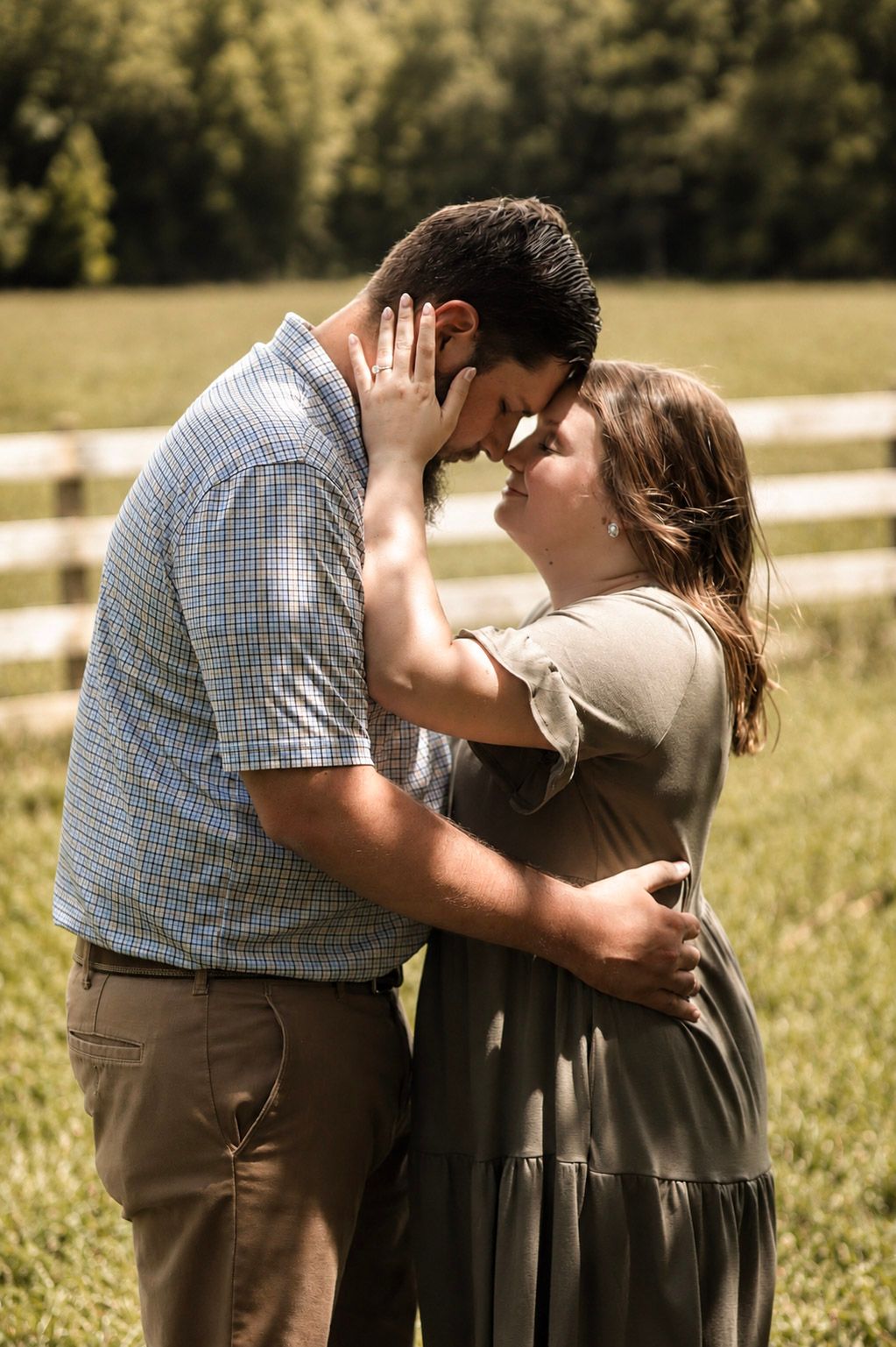 A couple shares a romantic forehead touch in a sunny green field beside a white wooden fence.