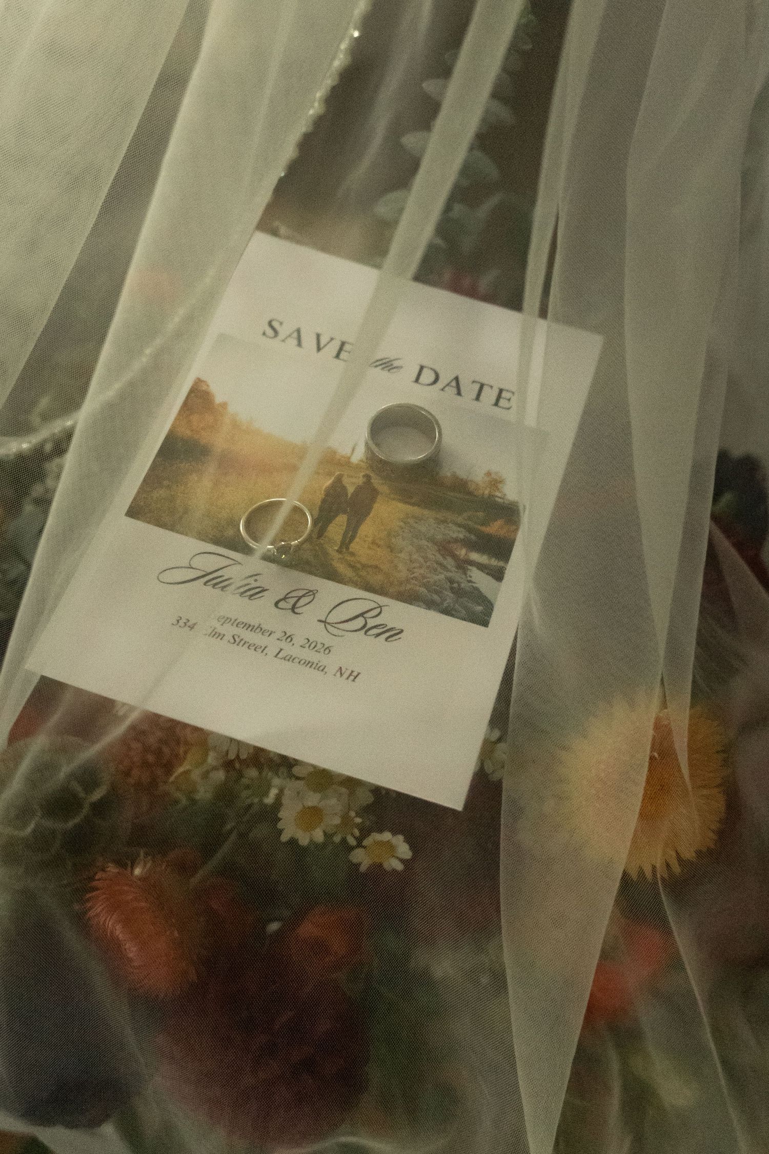 Save the date card with two wedding rings resting on top, surrounded by sheer veil fabric and colorful dried flowers.