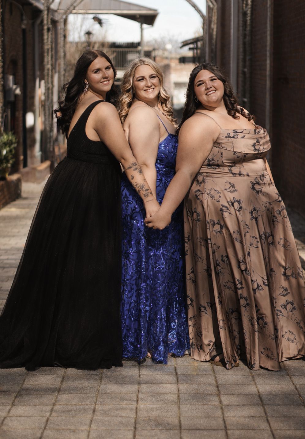 Three formal dresses in black, blue sequin, and floral print are showcased against a brick wall backdrop.