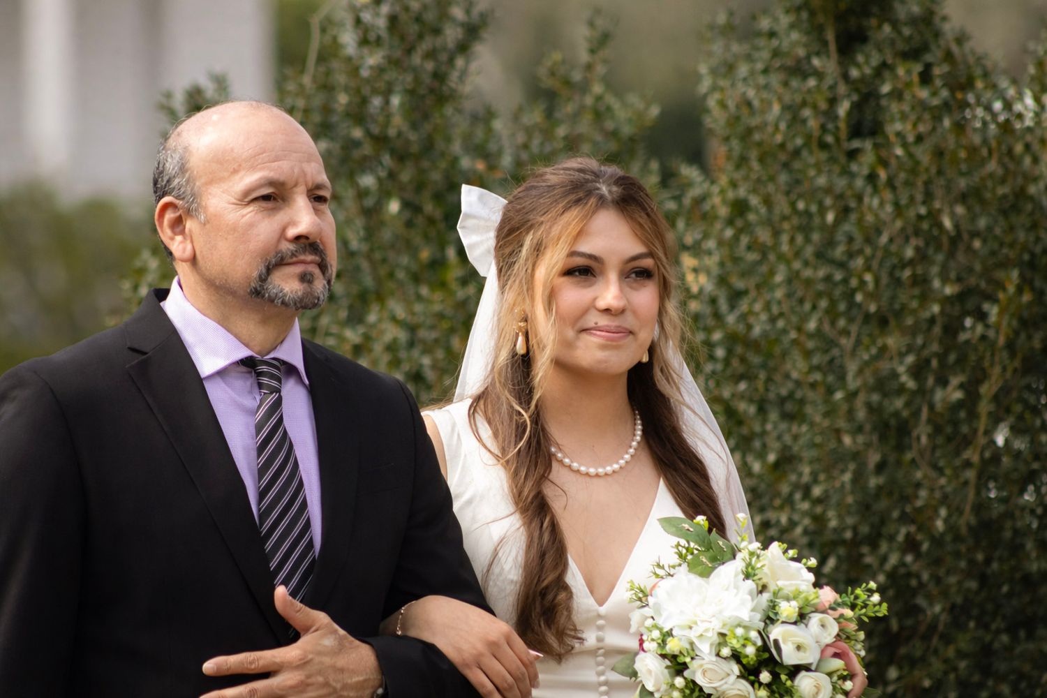 A wedding party poses outdoors in formal attire among green foliage on a sunny day.
