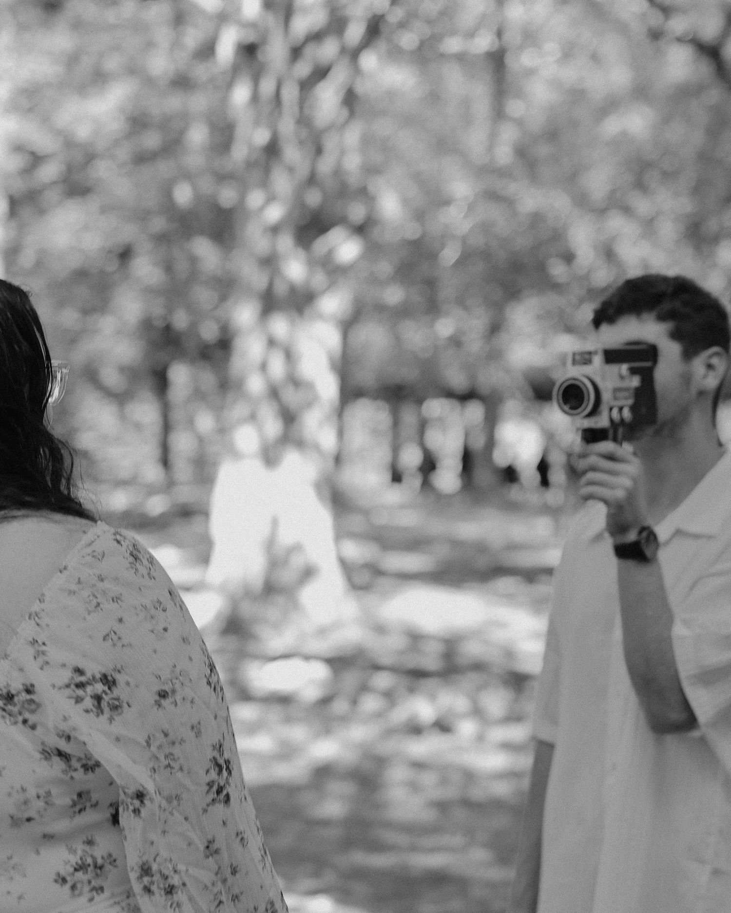 Black and white photo of a man filming a woman with a vintage camera outdoors among trees.