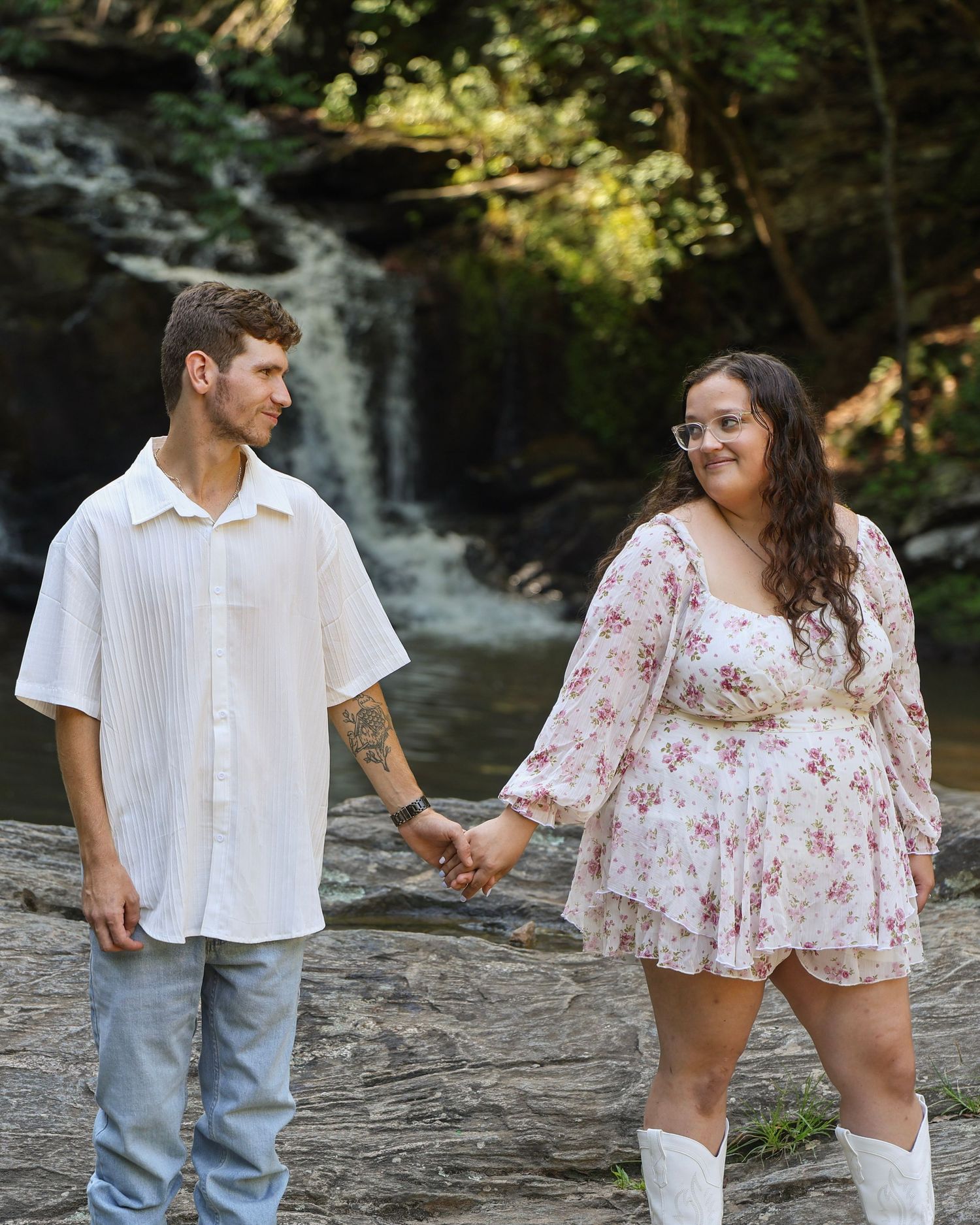 Couple holding hands in front of a waterfall, man in white shirt and jeans, woman in floral dress and white boots.