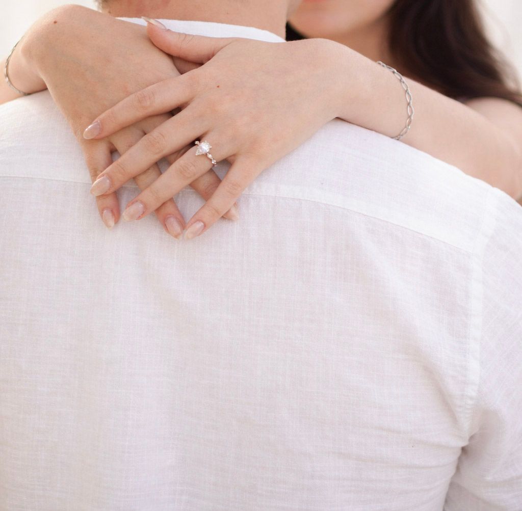 Close-up of hands with engagement ring resting on soft white bedding in natural light.