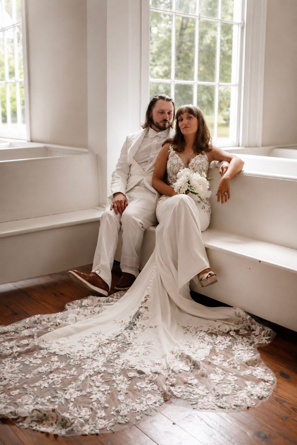 Bride and groom sitting together by window, bride wearing lace train wedding dress.