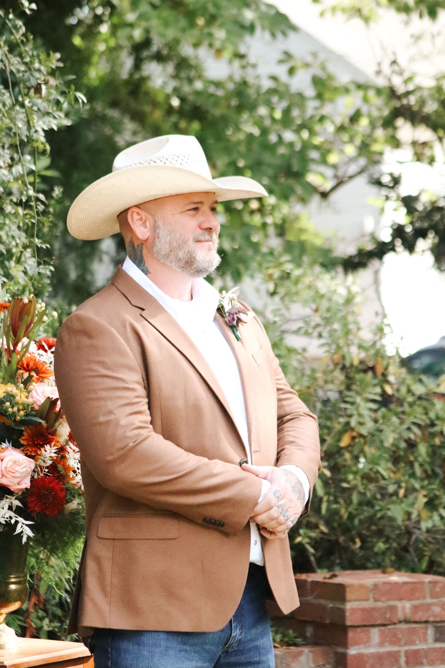 Man in brown blazer and cowboy hat poses against brick wall with autumn floral decorations.