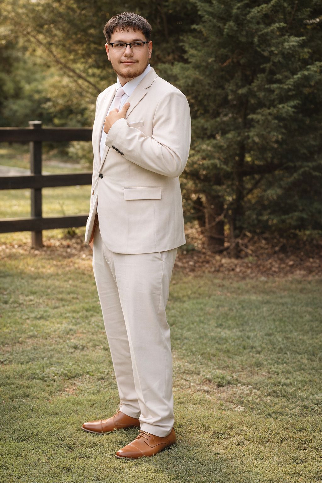Young man in beige suit and brown dress shoes poses outdoors near wooden fence and evergreen trees.