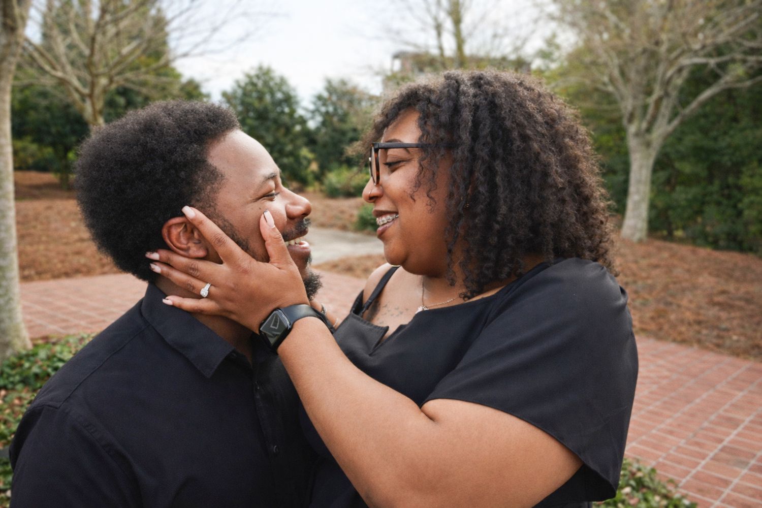 Two people share a tender embrace outdoors in a garden setting with natural lighting and greenery in the background.