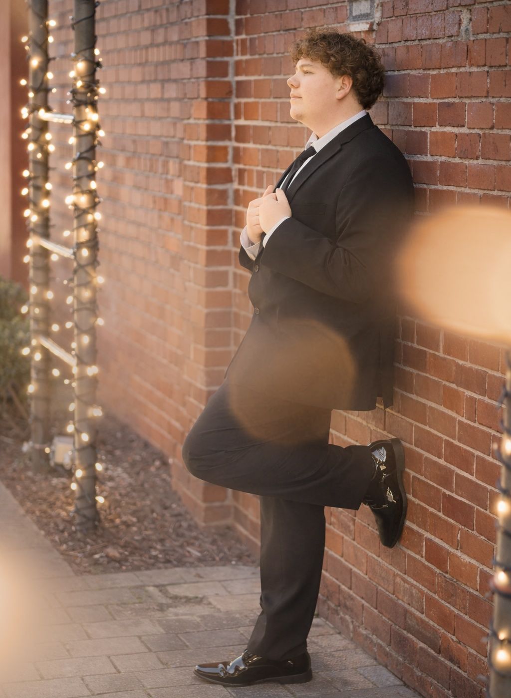 A sequence of formal photos against a brick wall with string lights showing someone in a black suit striking poses.