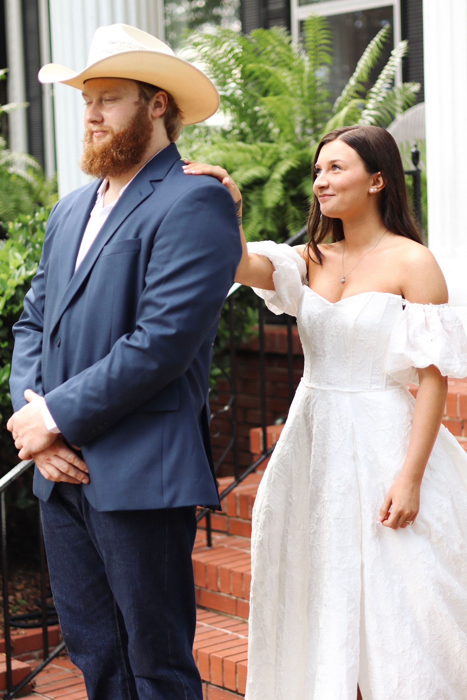A couple in wedding attire poses near brick columns with lush greenery in the background.