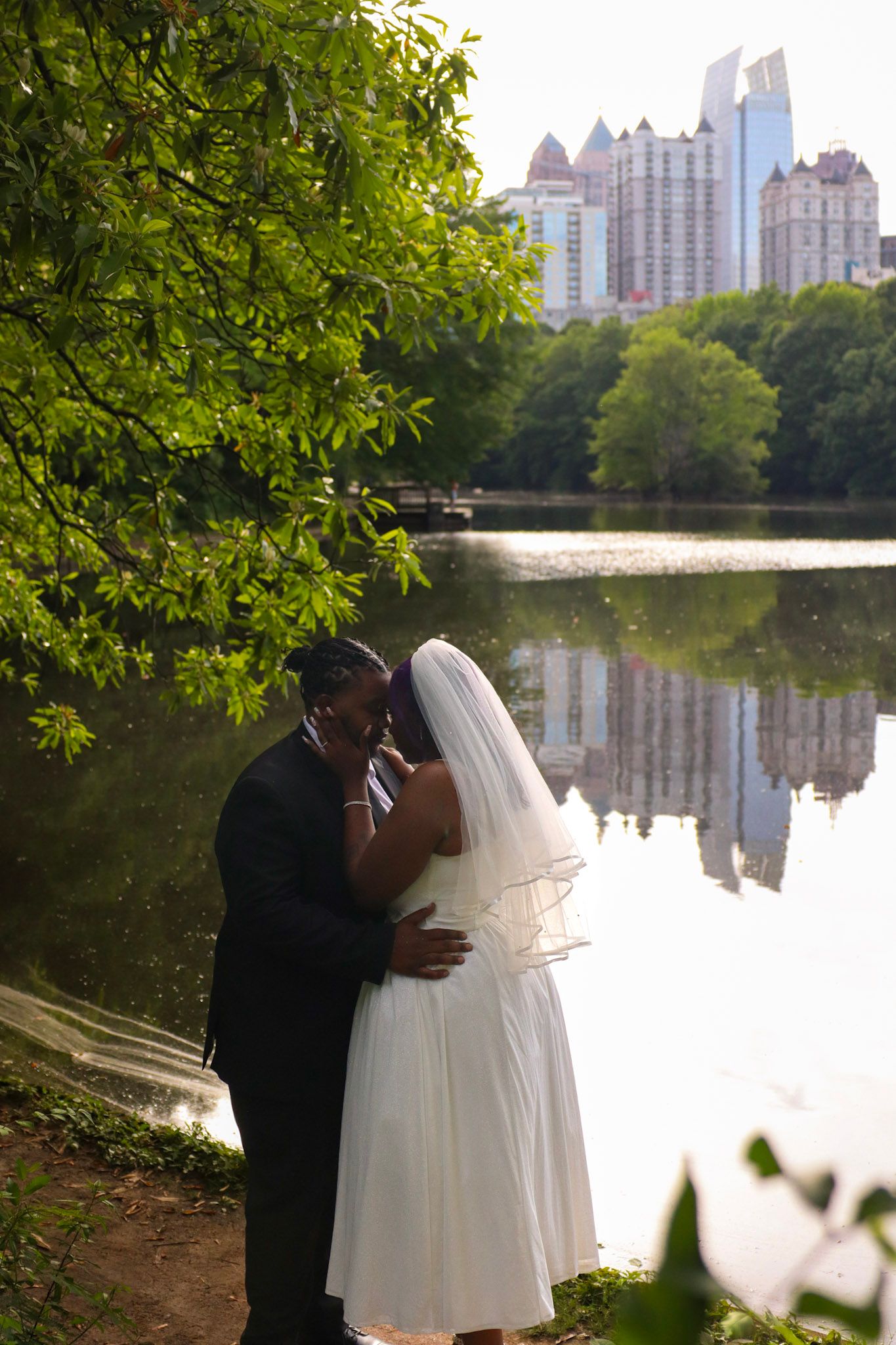 African American bride and groom share a romantic embrace by a reflective lake with city skyline backdrop.