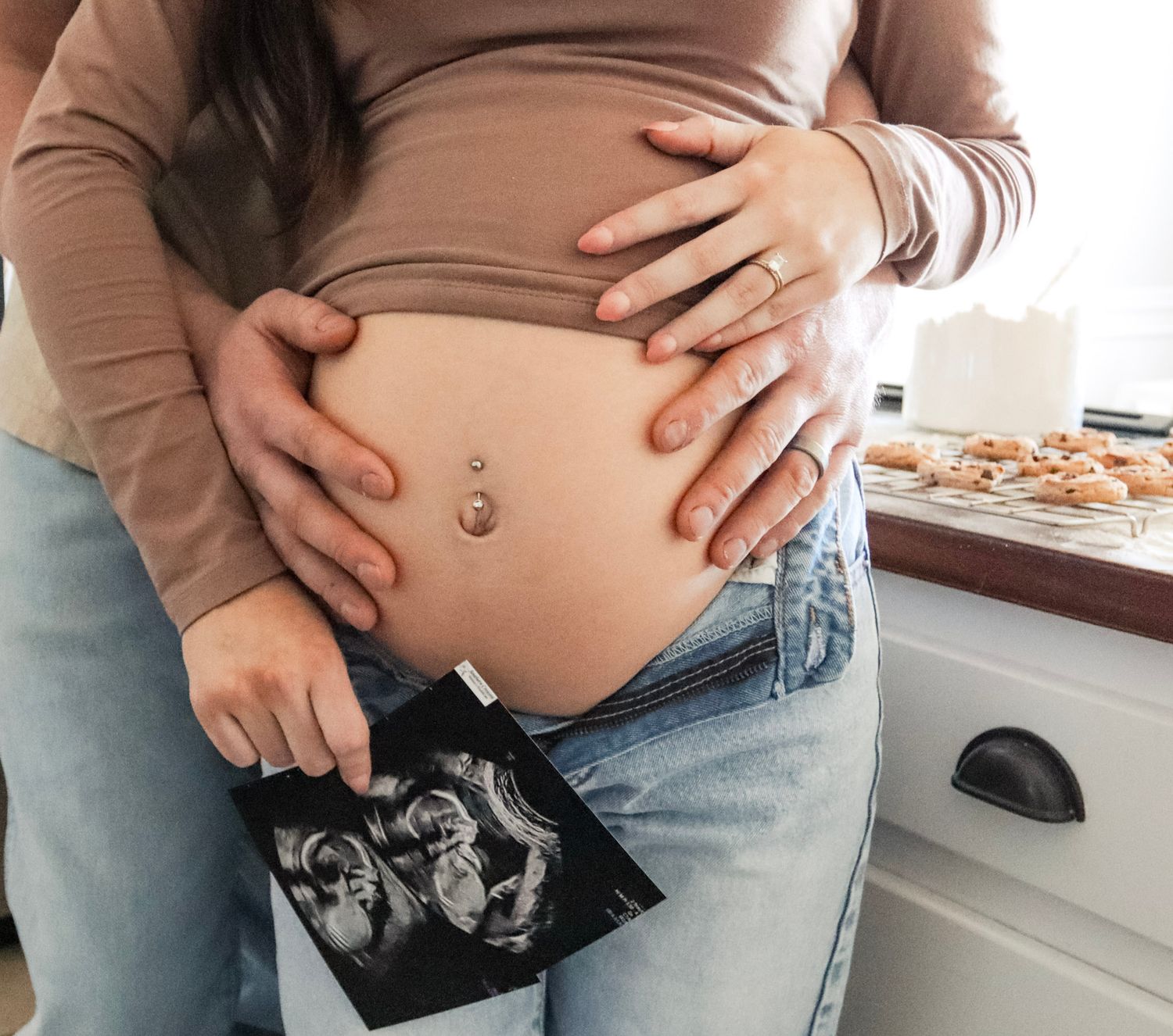 Pregnant woman holding ultrasound photo while partner embraces baby bump in cozy kitchen setting.