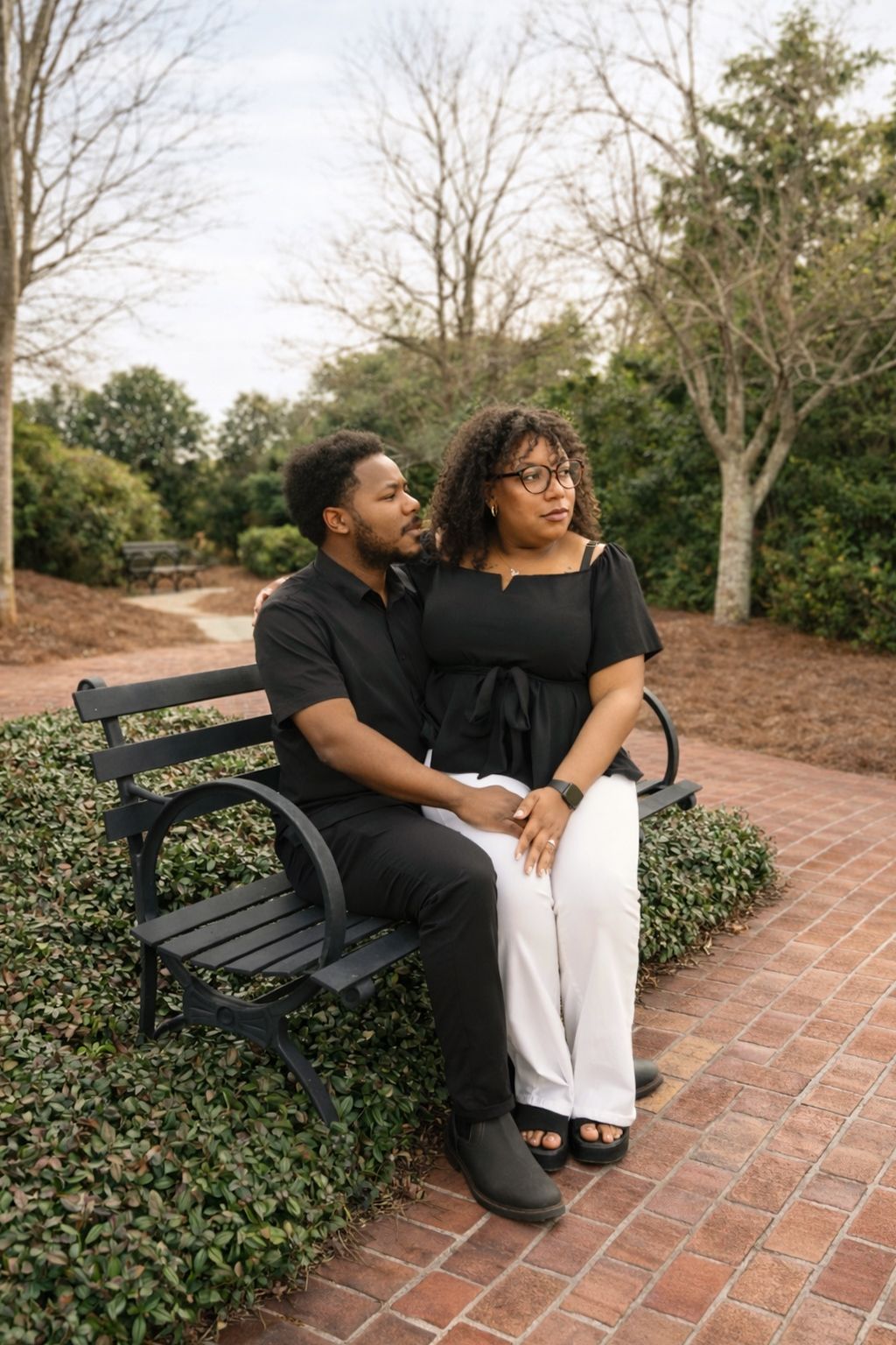 A couple sits together on a park bench along a brick pathway surrounded by greenery on a peaceful afternoon.