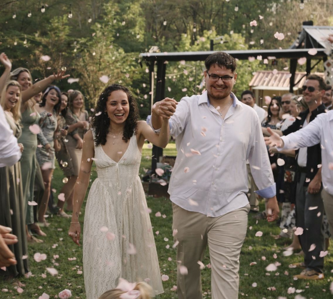 A joyful couple walks through a shower of rose petals at their outdoor wedding ceremony surrounded by smiling guests.