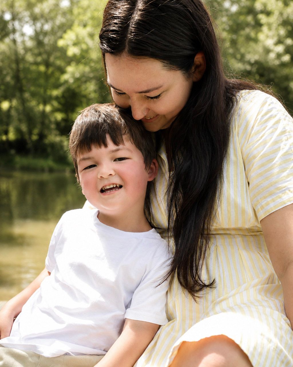 A smiling mother in a yellow striped dress cuddles her laughing young son outdoors near a serene pond.