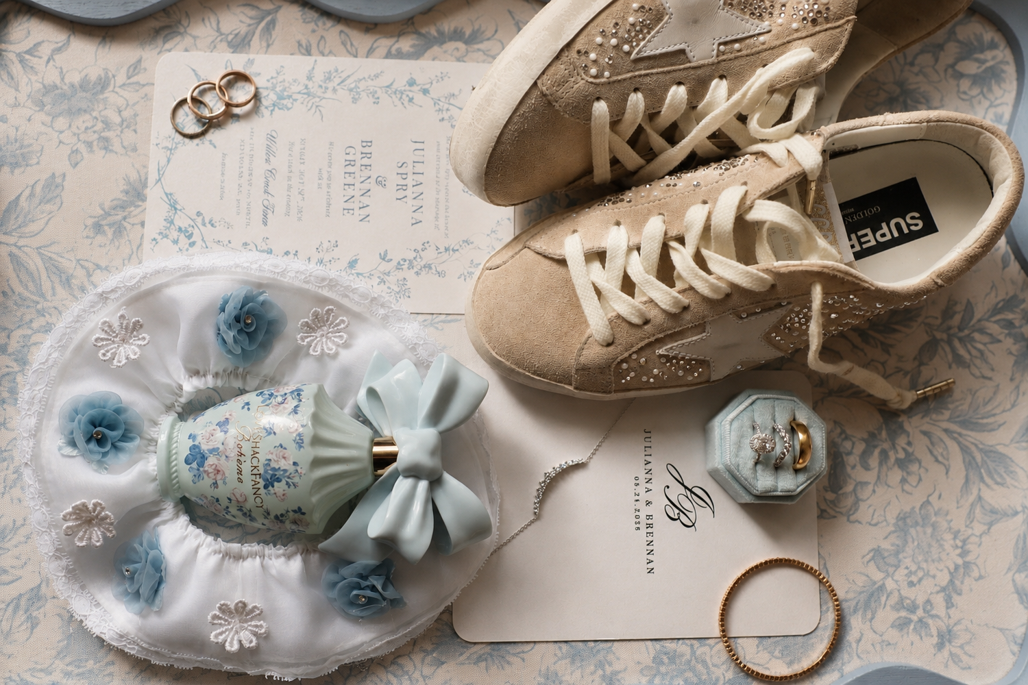 Flat lay of wedding details featuring golden sneakers, blue floral accessories, rings, and elegant stationery on a floral backdrop.