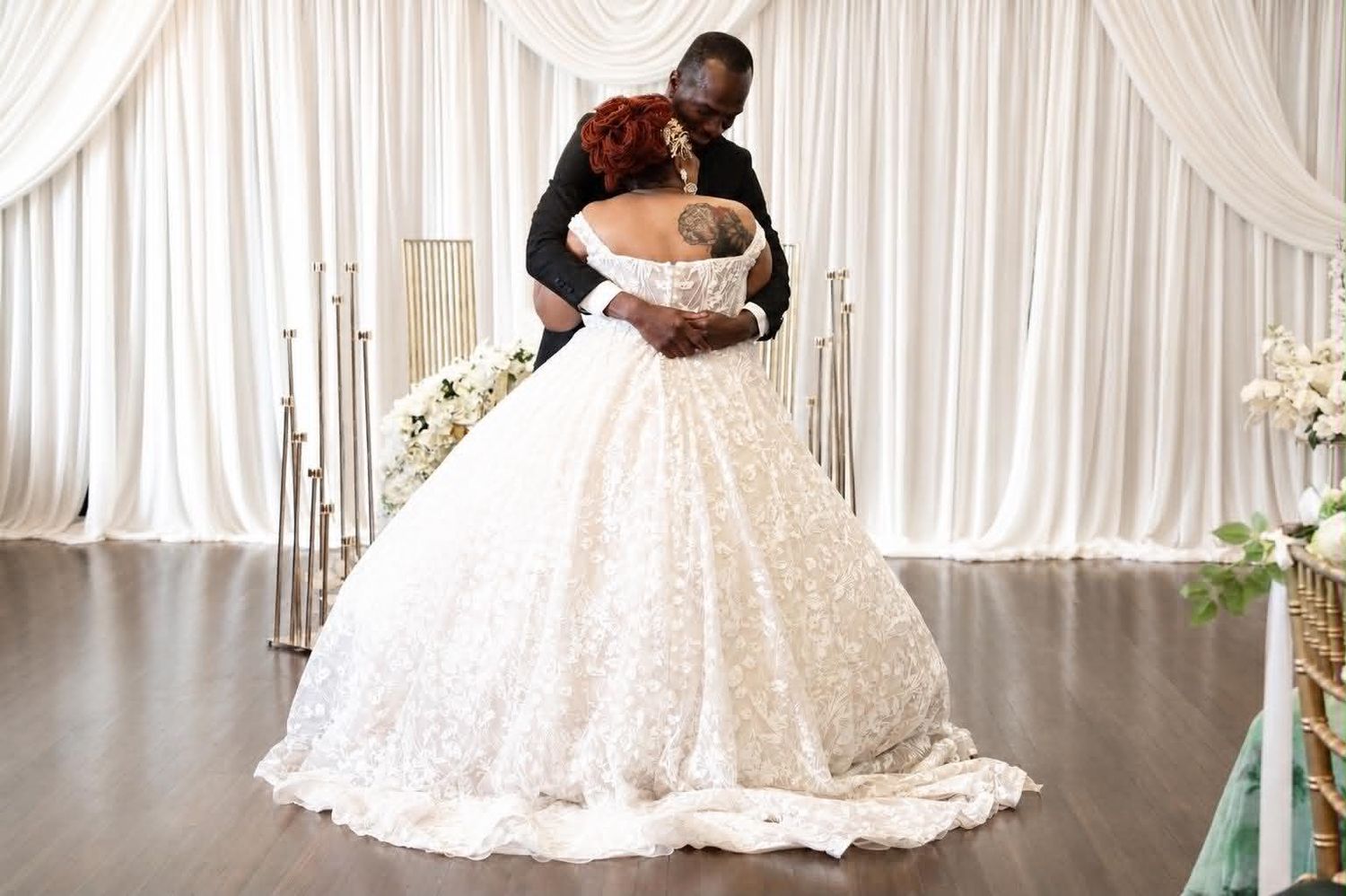 Couple sharing first dance at wedding reception, bride in elegant white ballgown, groom in black suit, draped white backdrop.