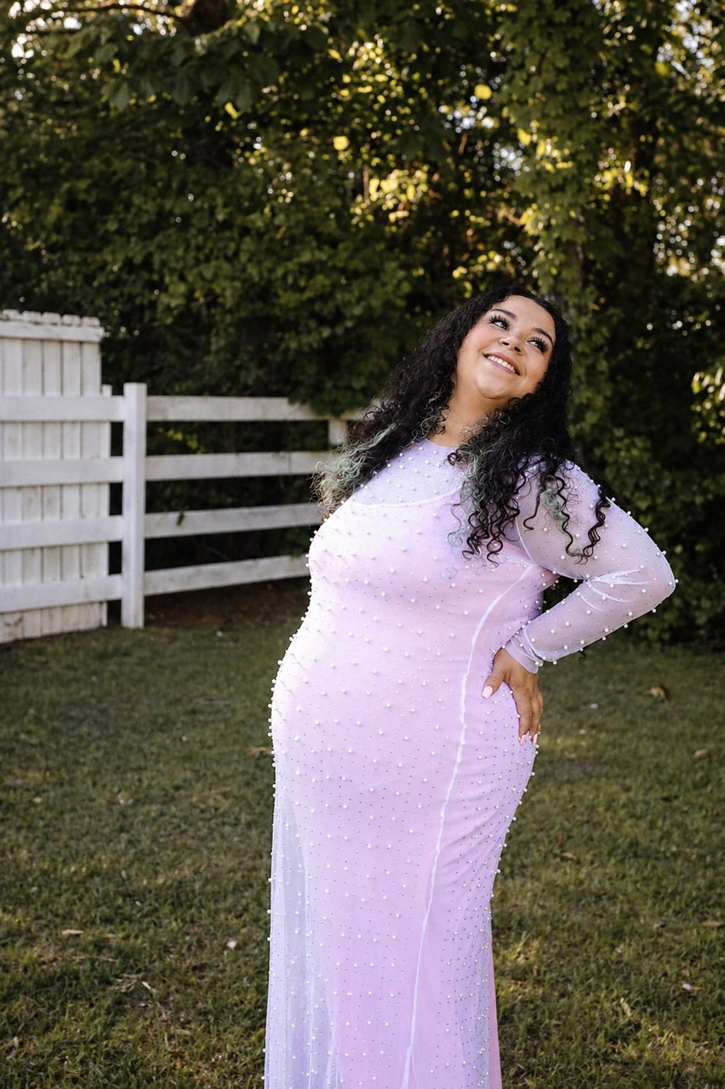A woman in a white beaded long-sleeve gown poses outdoors near a white fence and green trees.