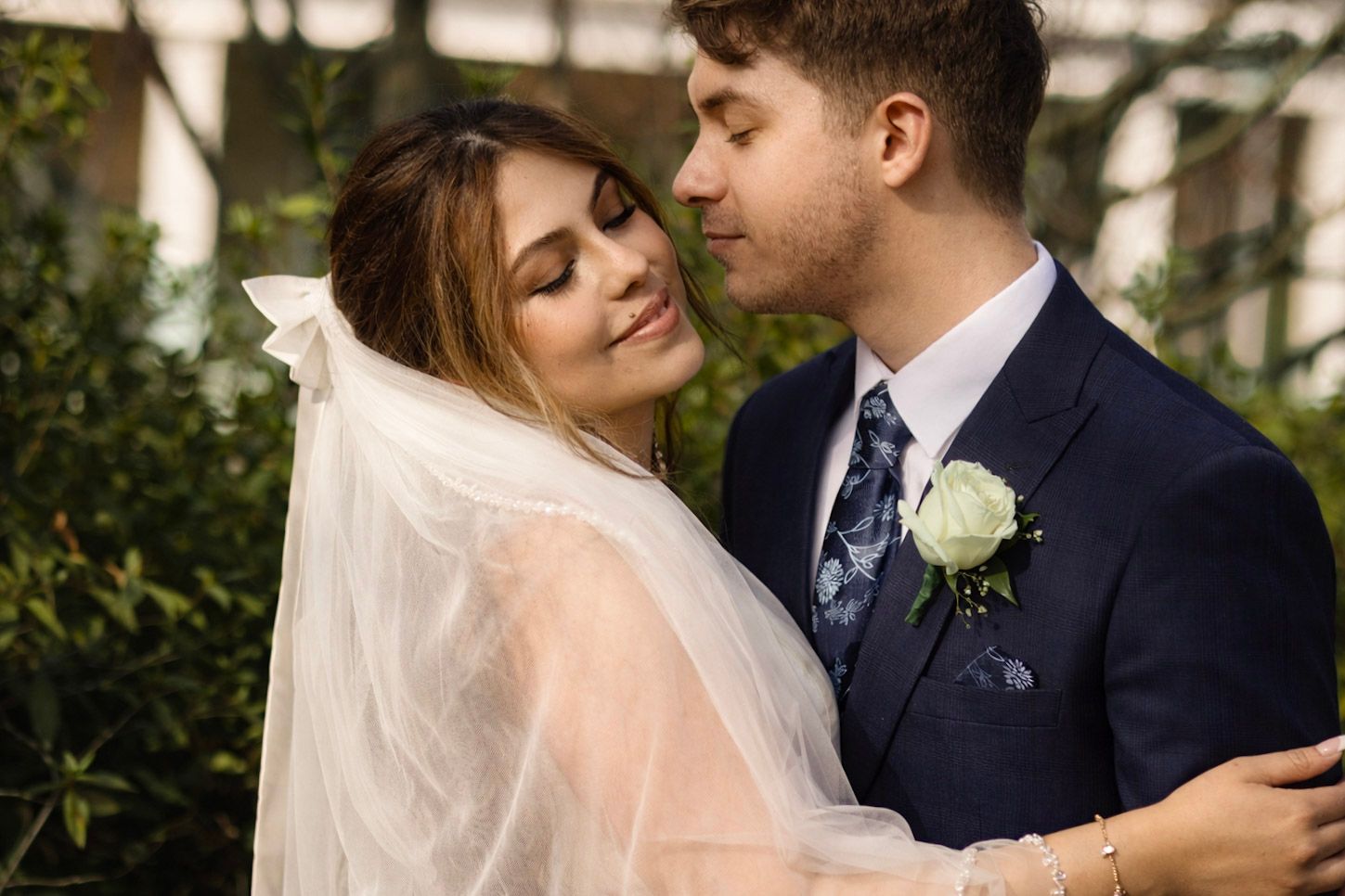 A bride and groom share an intimate moment outdoors, with her wearing a flowing veil and him in a navy suit with boutonniere.