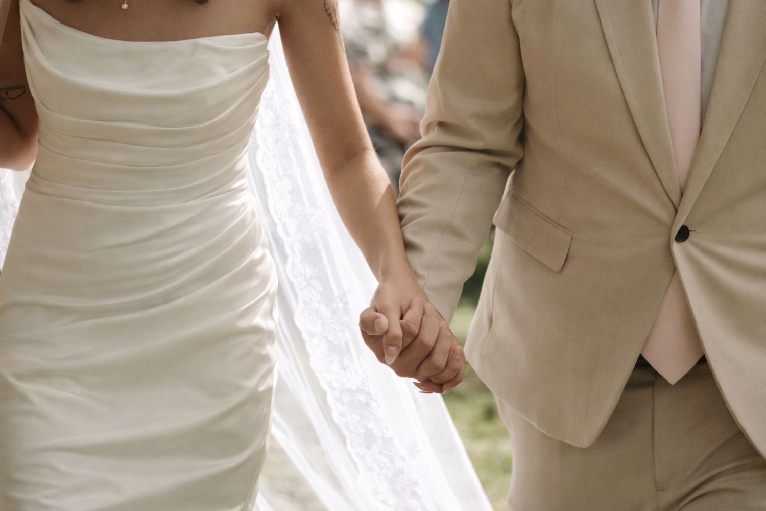 Bride in white strapless gown and groom in beige suit holding hands at outdoor wedding ceremony.