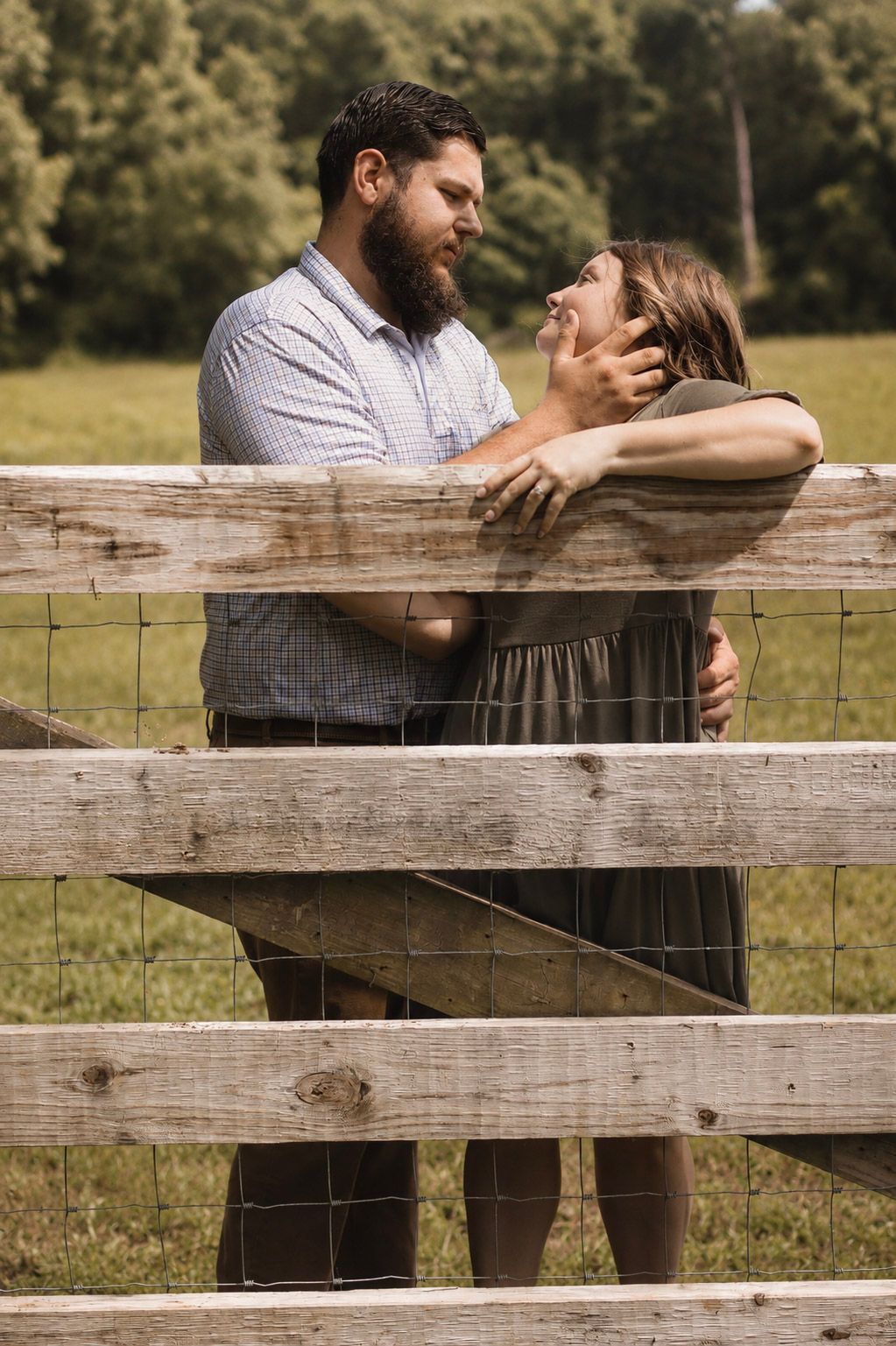 A bearded man and smiling woman share a romantic moment beside a rustic wooden farm fence in a green field.