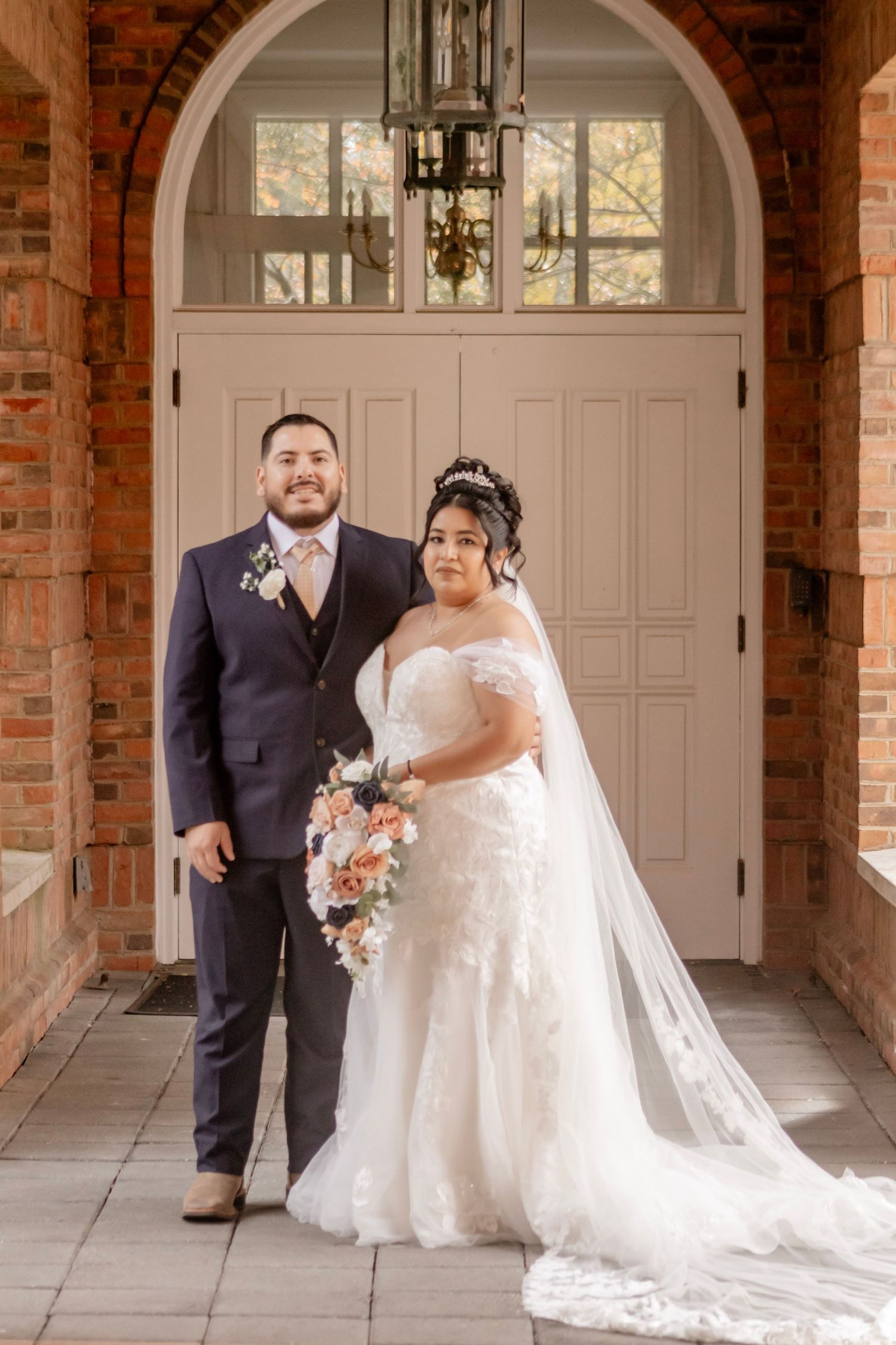 Bride and groom pose together in an elegant brick archway with white doors and natural lighting.