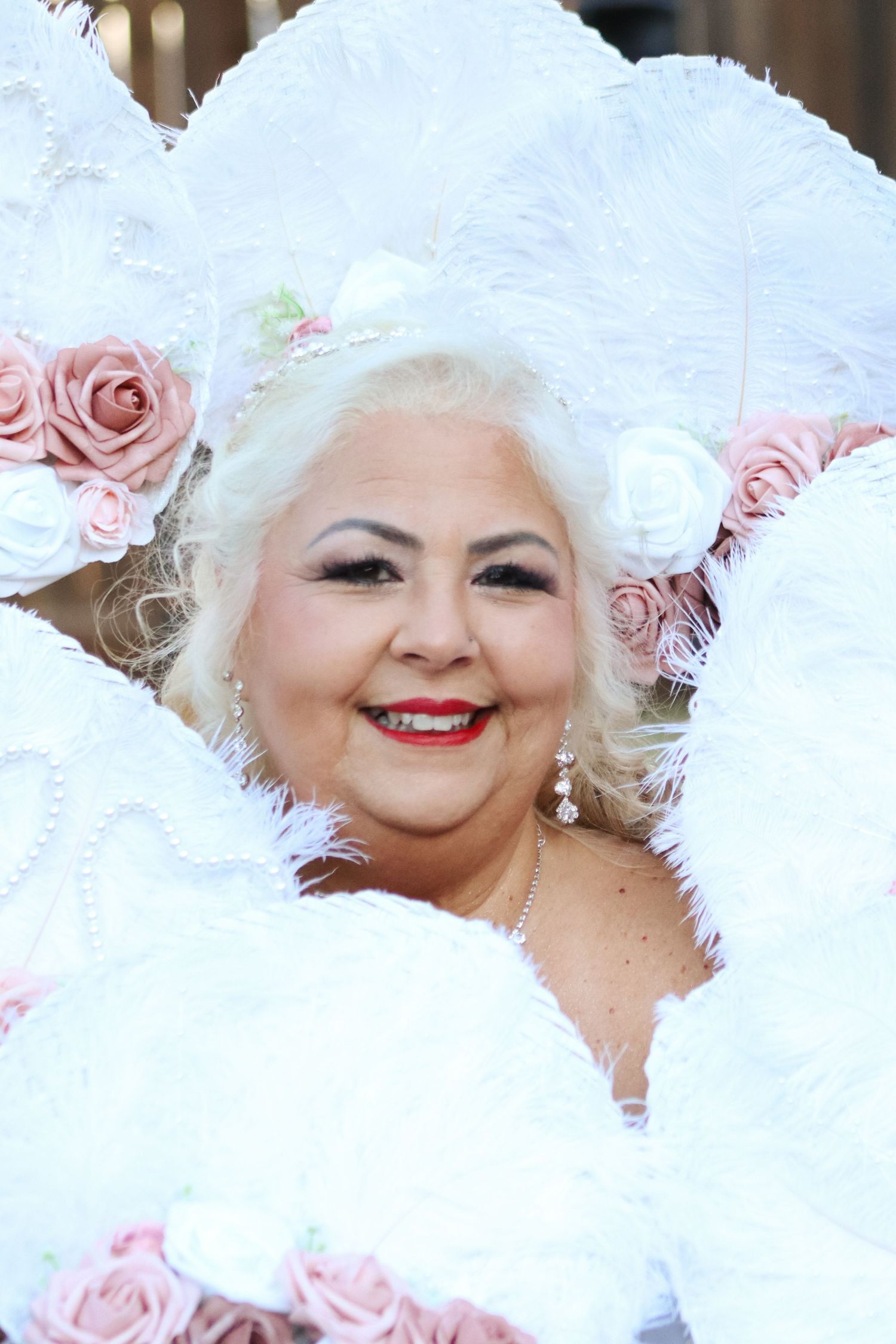 Bride wearing ornate white feathered hat with pink roses and red lipstick smiles joyfully.