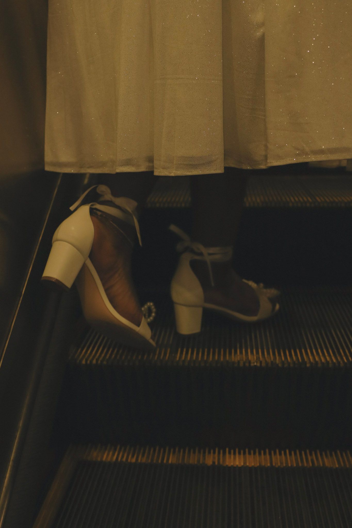 Close-up of white block-heel sandals on an escalator with a flowing white dress in the background.