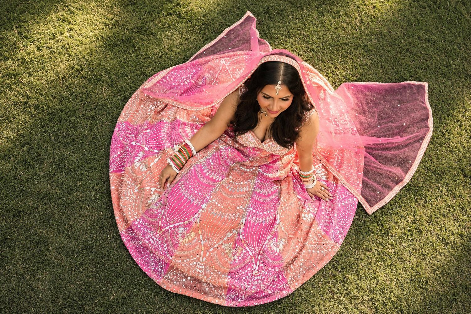Woman in stunning pink embroidered lehenga choli sitting on grass, photographed from above, showcasing full skirt spread.