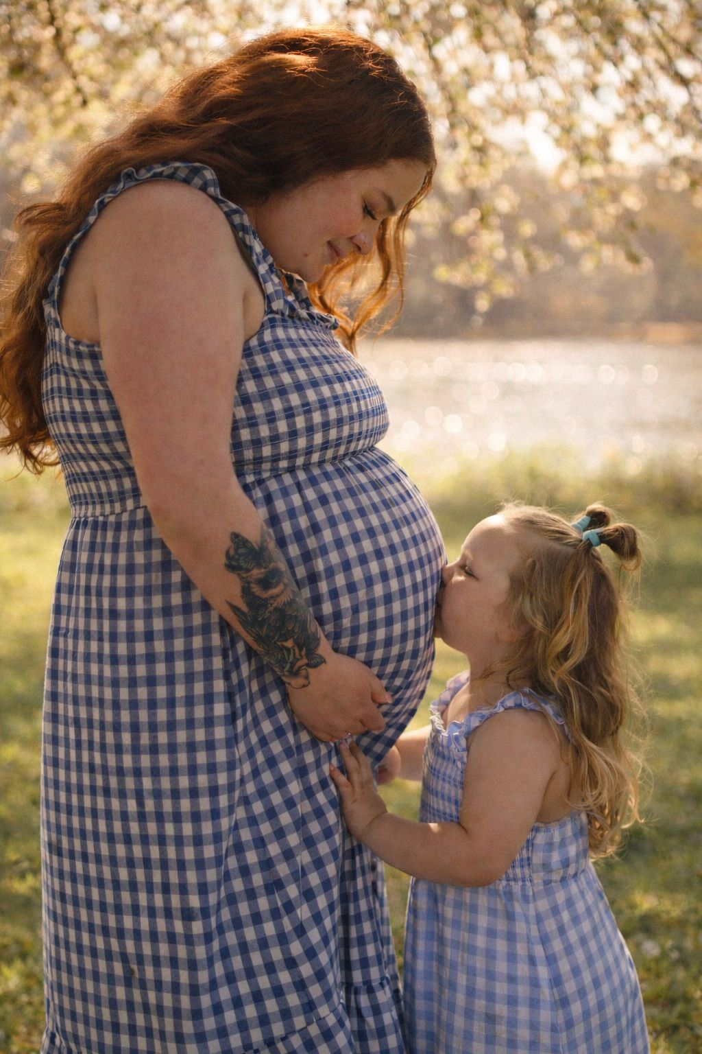 Pregnant woman and little girl in matching blue gingham dresses share a tender moment outdoors in golden sunlight.