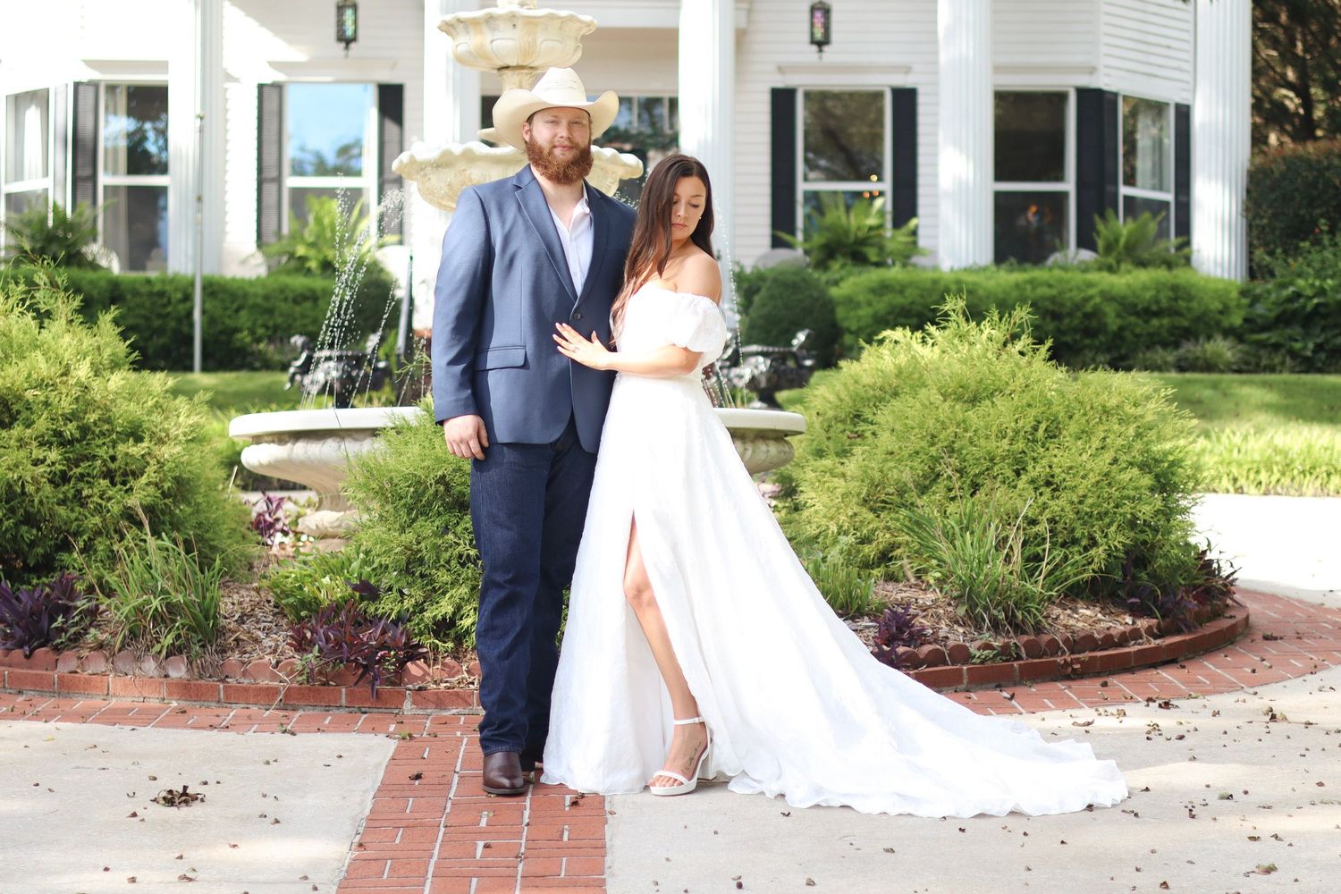 Wedding couple poses on brick pathway in front of white columned building with landscaped gardens.