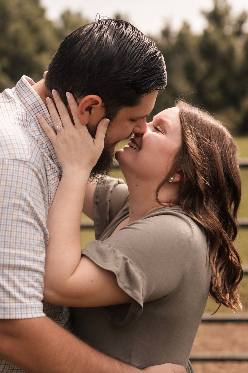 A couple shares a romantic kiss outdoors, with the woman smiling joyfully as they embrace in a lush green setting.