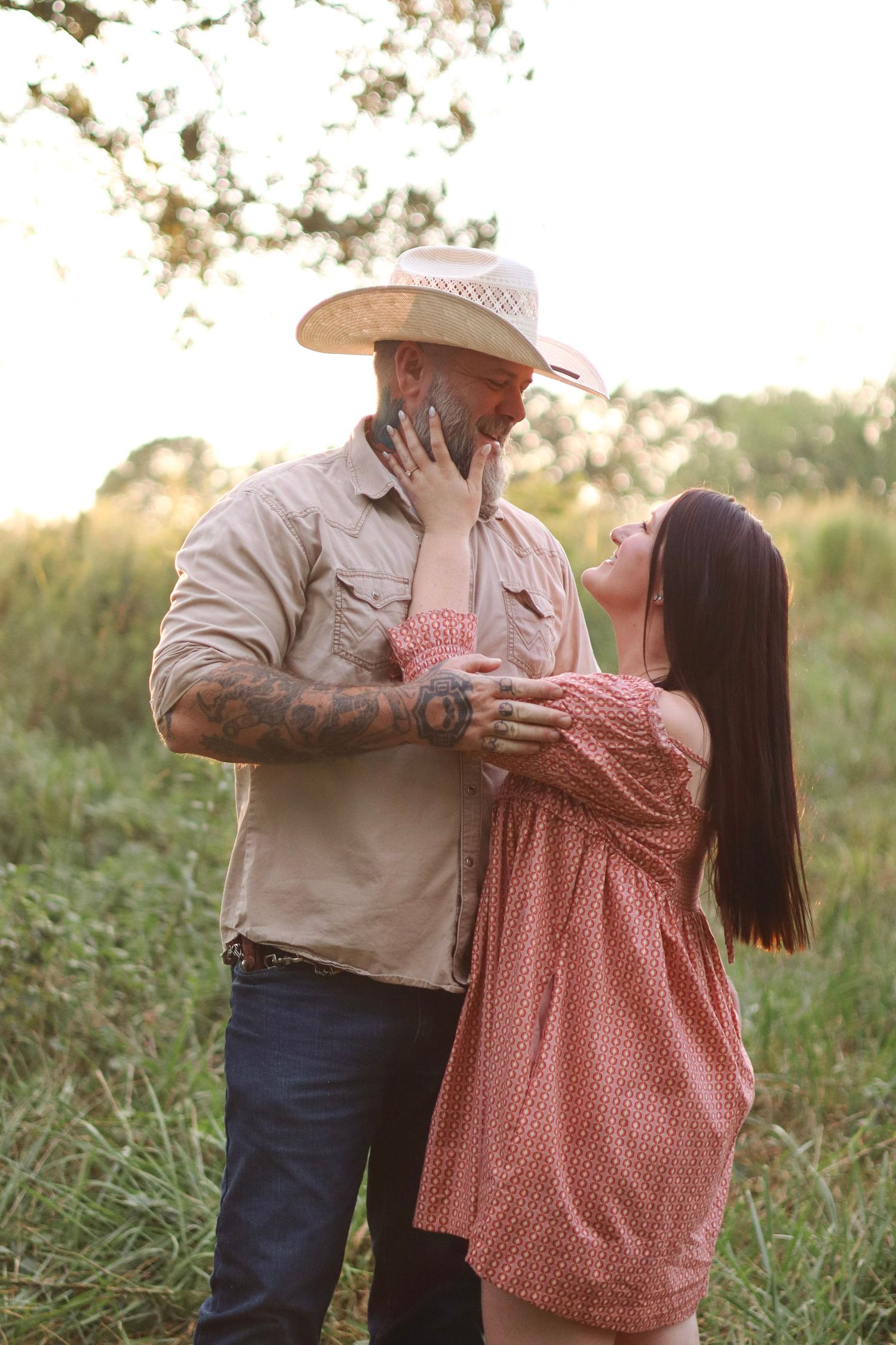 A couple embraces in a golden field during sunset, with the person wearing a cowboy hat and tan shirt.