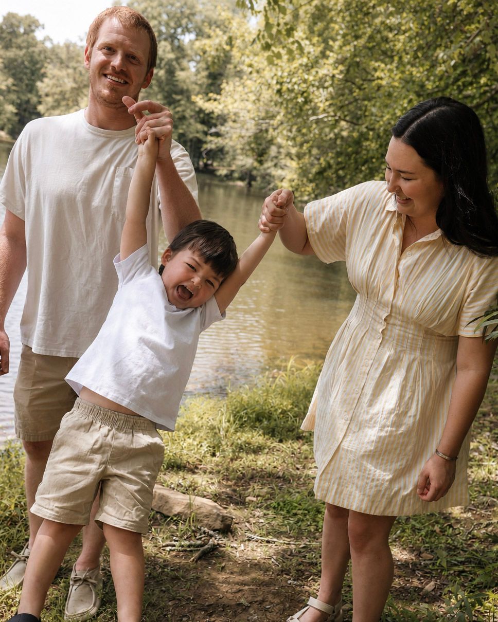 A happy family of three swinging their toddler by a sunlit riverside park on a warm summer day.