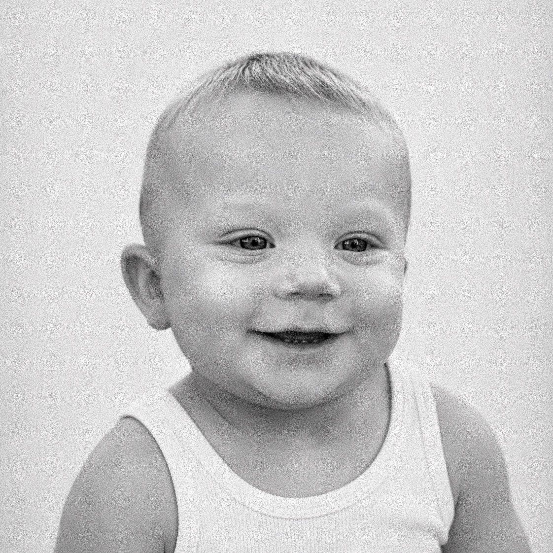 Black and white portrait of a happy smiling baby wearing a white tank top against a light background.