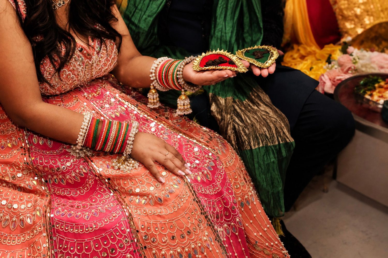 Close-up of bride in pink embroidered lehenga with green bangles holding floral kalira at Indian wedding ceremony.