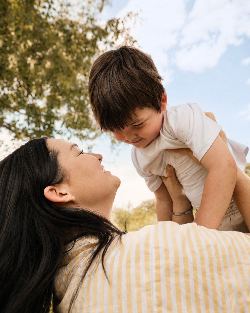 A mother joyfully lifts her smiling toddler in the air outdoors, sharing a tender moment under a blue sky.