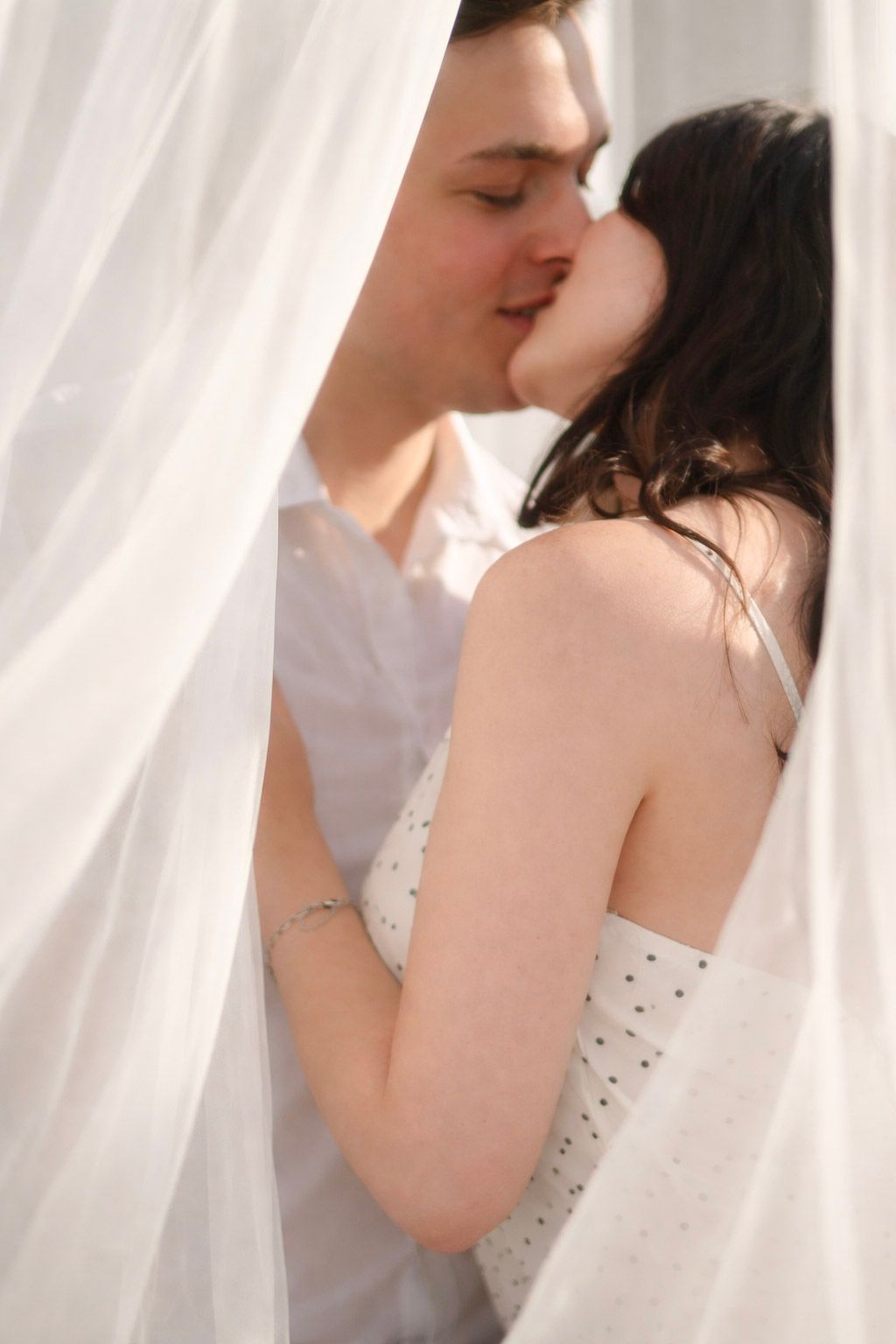 A romantic couple shares an intimate kiss behind sheer white curtains in a bright, dreamy setting.