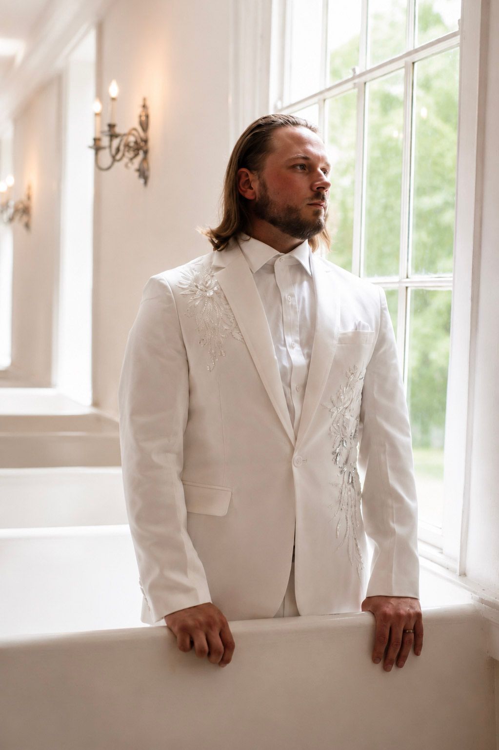Groom in white embroidered suit jacket standing by large window on wedding day.