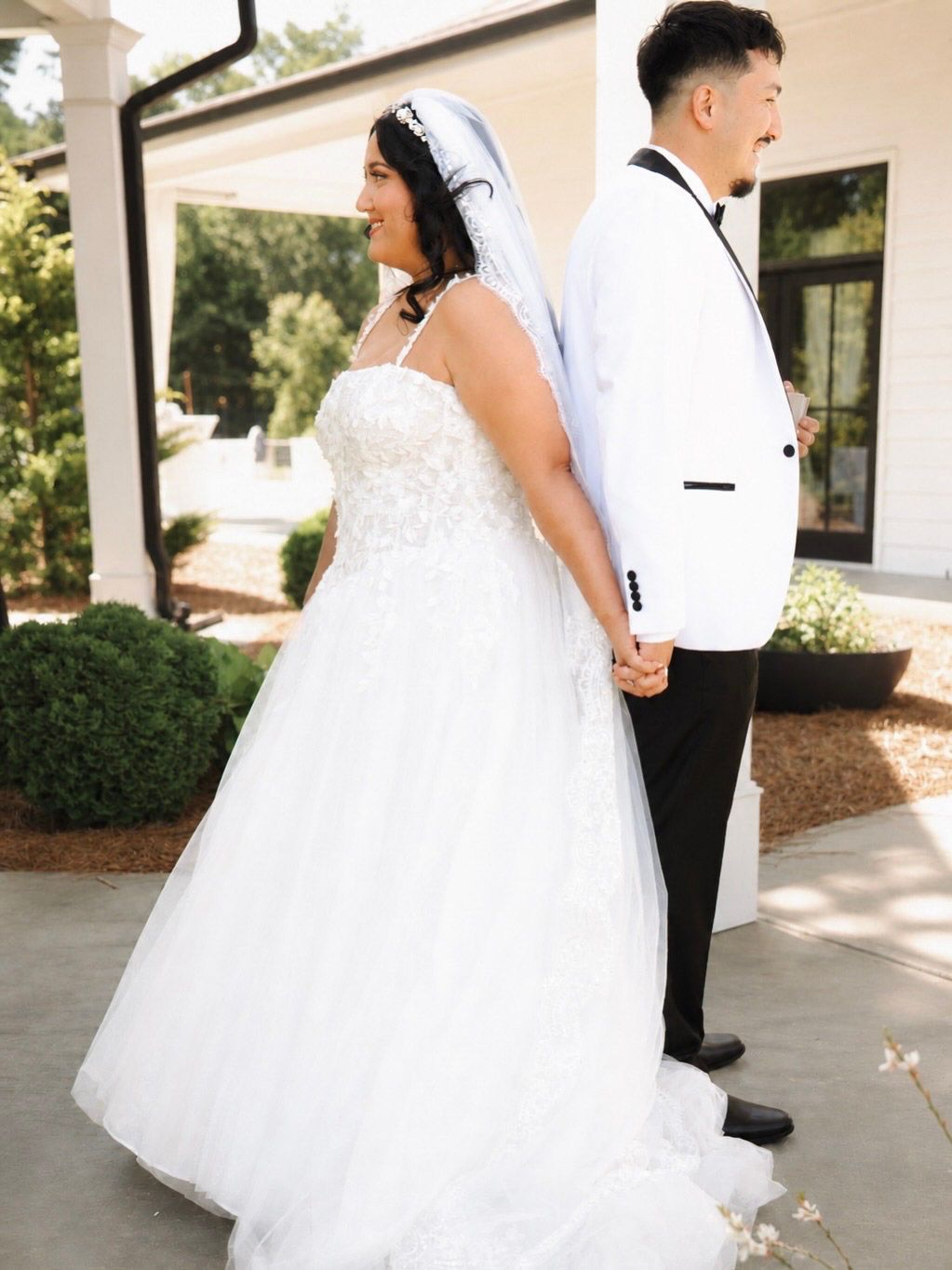 Bride in white ballgown and veil holds hands with groom in white tuxedo jacket outdoors at wedding venue.