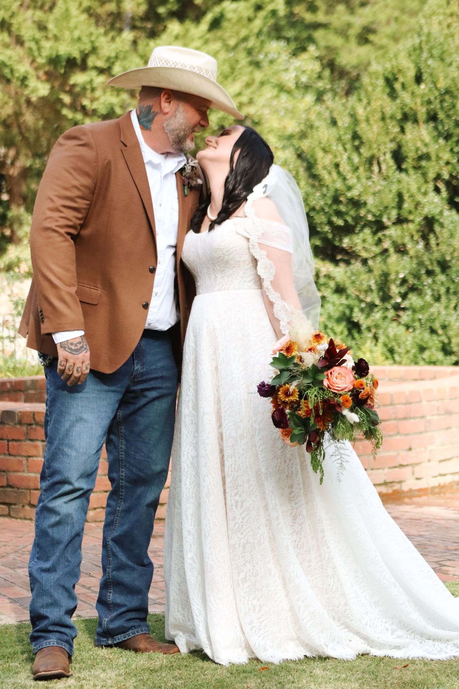 Wedding couple poses together outdoors, with the bride in white lace and groom in brown jacket and cowboy hat.