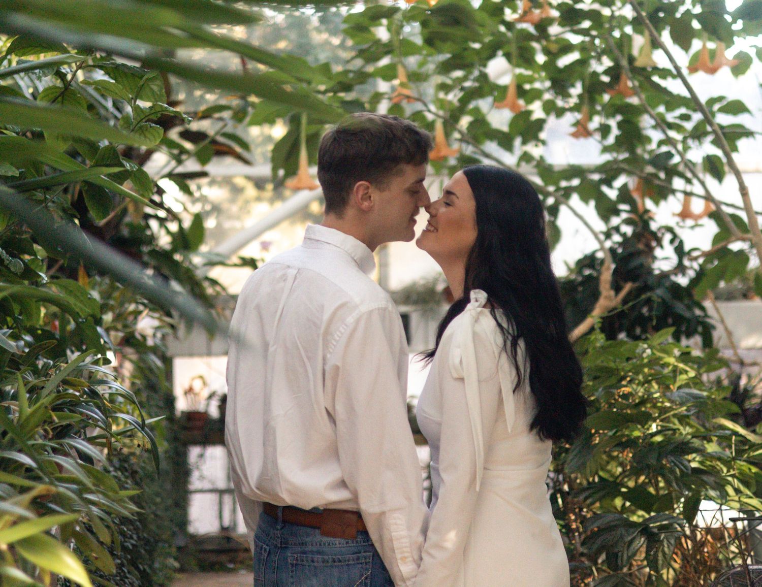 A couple shares a romantic moment under lush green foliage in an outdoor garden setting.