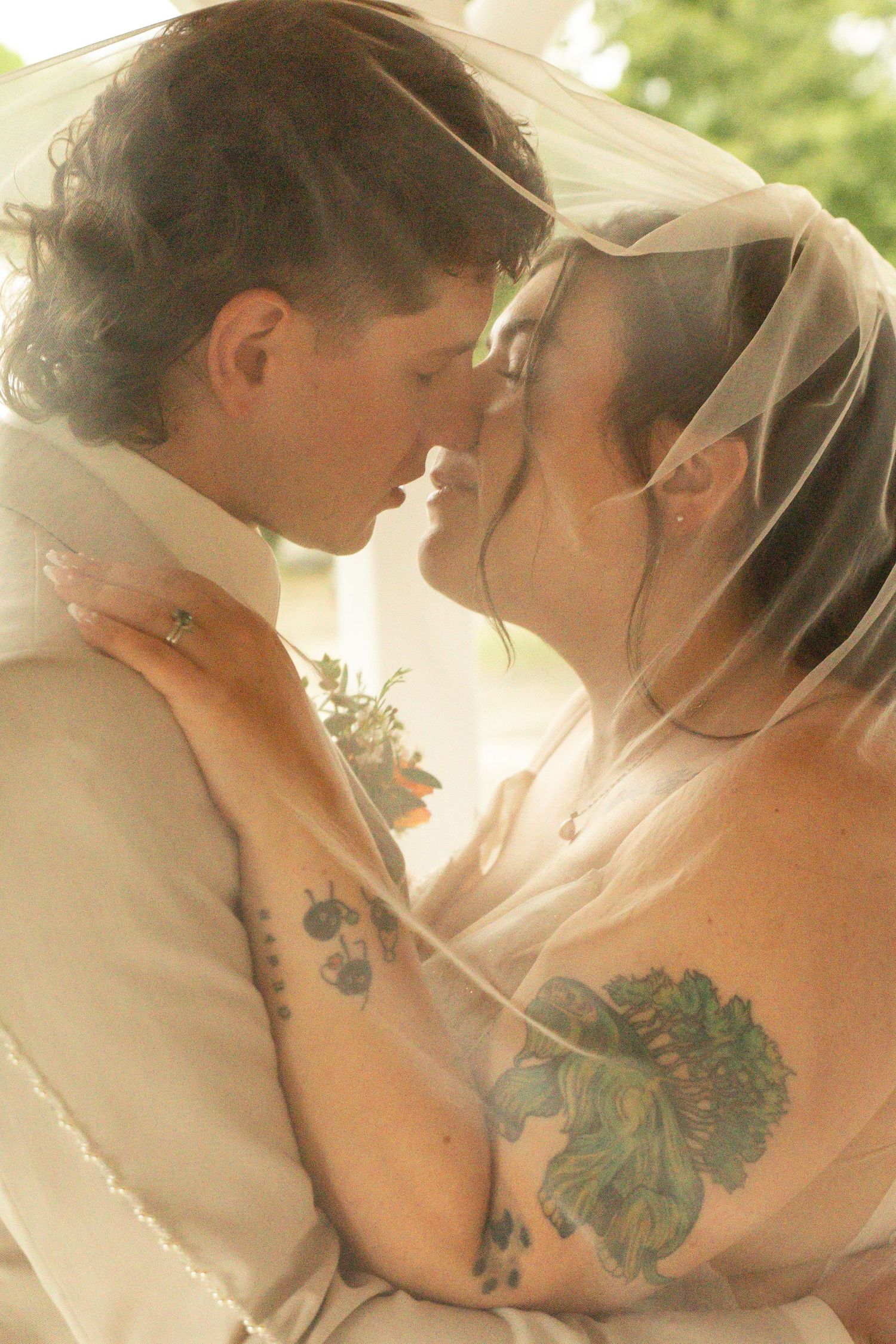 Tattooed bride and groom in a tender moment about to kiss beneath a sheer wedding veil, with a floral boutonniere and an outdoor background.